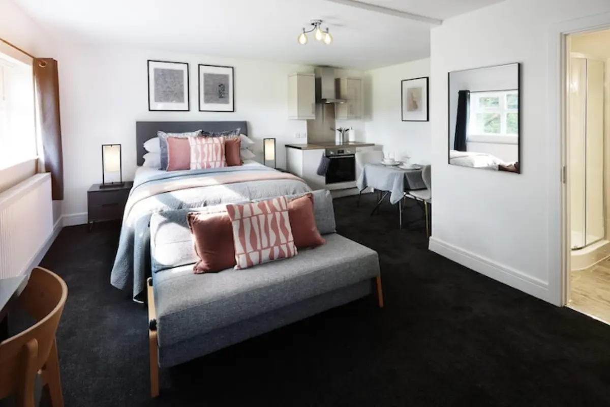 Modern self-contained student Modern self-contained student studio apartment interior featuring a double bed, integrated kitchenette, small dining table, small sofa, and an open doorway leading to a private ensuite bathroom